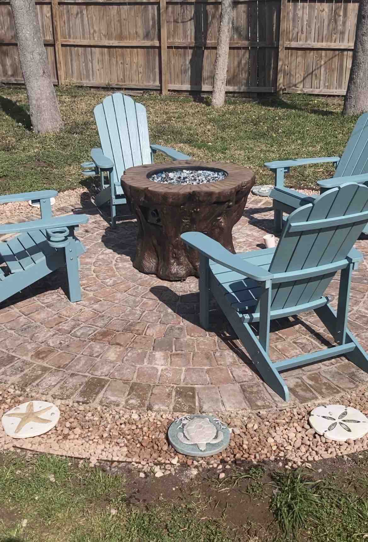 A circular seating area is visible, featuring four blue Adirondack chairs arranged around a fire pit made of natural wood. The brick patio is surrounded by decorative stones, enhancing the outdoor ambiance. Green grass and wooden fencing are present in the background.