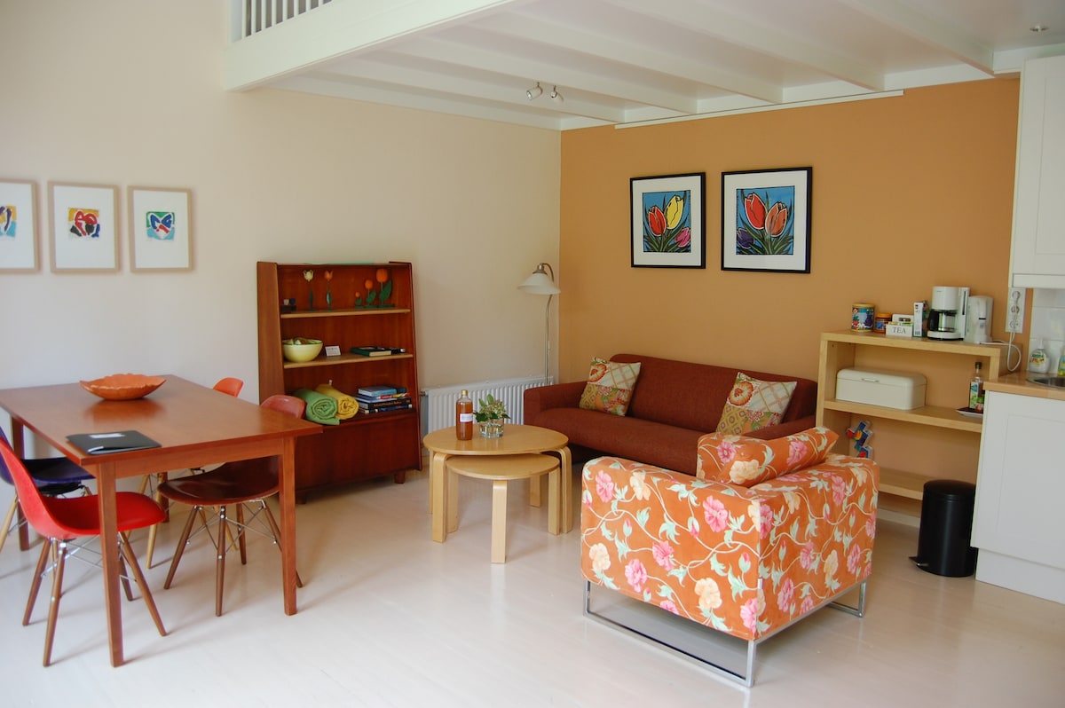 A bright living space features a dining table with four chairs and a cozy seating area with a floral-patterned couch. Two framed artworks adorn the wall, complemented by a wooden shelving unit displaying various items. A kitchenette area is visible, enhancing functionality.