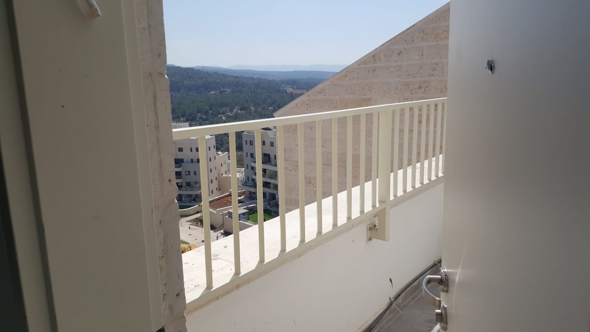 A balcony view is presented, featuring a railing that overlooks the surrounding area. The urban landscape includes a mix of buildings and greenery extending into the distance, with hills outlined against the sky. The setting captures a sense of openness and connection to the surroundings.