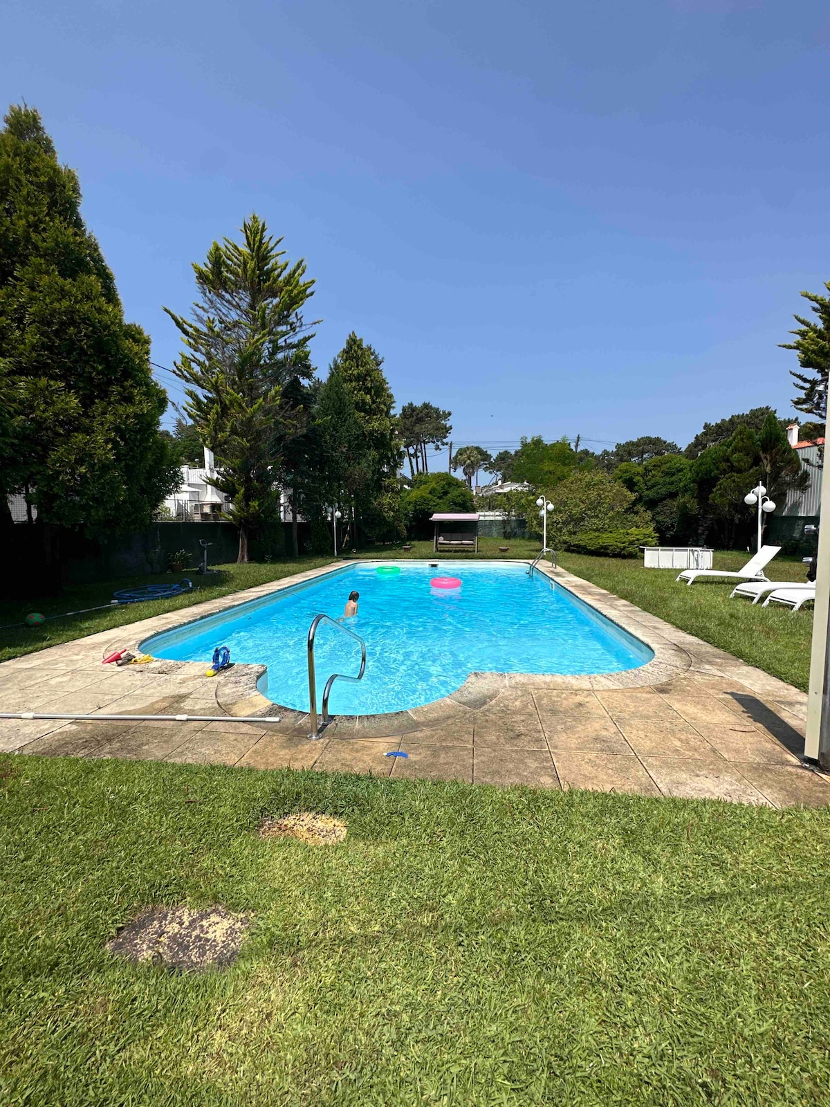 A well-maintained swimming pool is surrounded by a grassy area, with several lounge chairs positioned nearby. Colorful pool floats are seen in the clear blue water. Lush trees provide a backdrop, adding to the outdoor setting's natural ambiance.