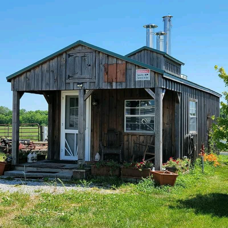 A rustic cabin features a wooden exterior with a tin roof. Brightly colored flower pots line the pathway to the front door, which has a glass panel. Large windows allow light in, offering views of the surrounding green pastures.