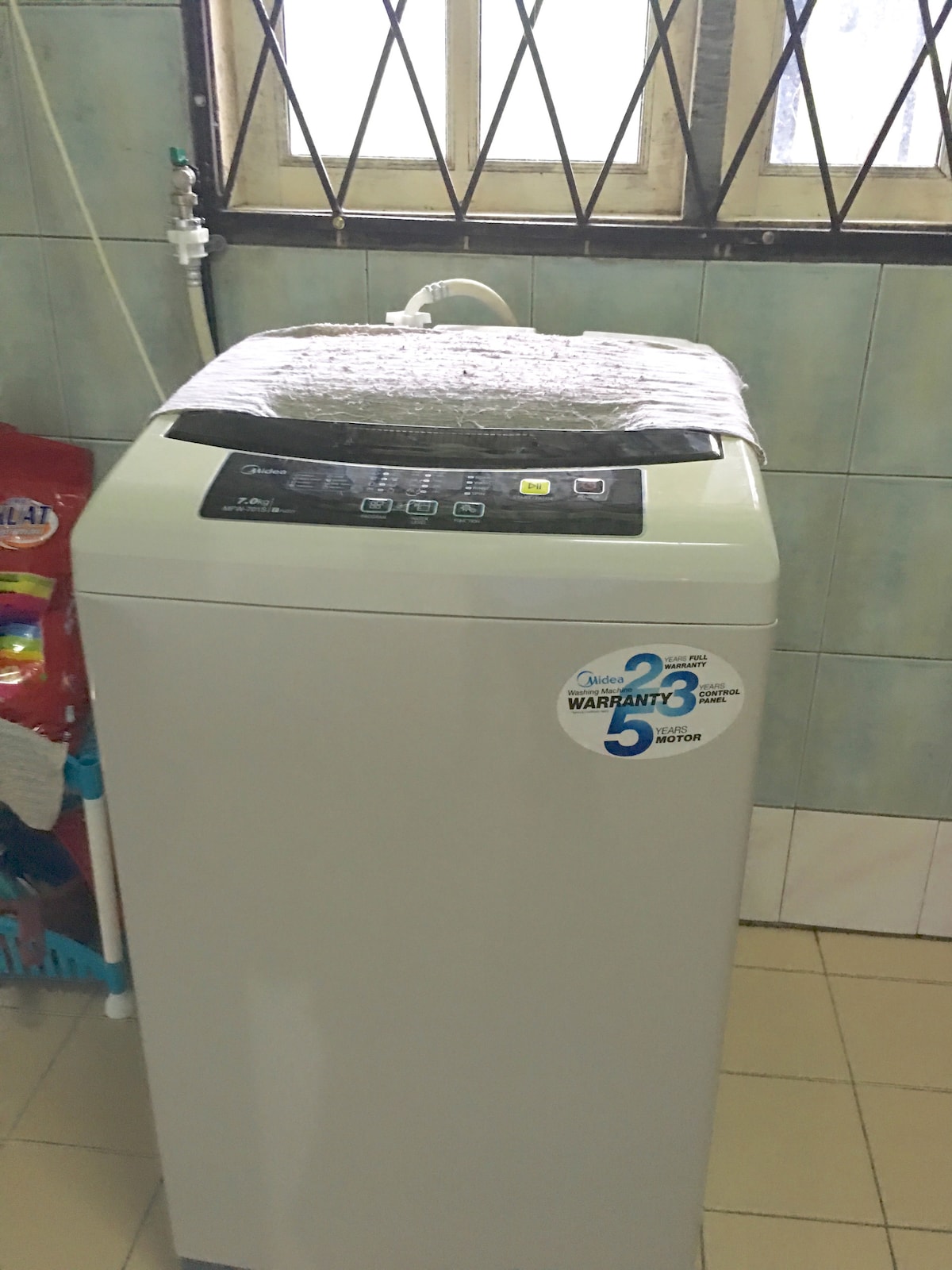 A modern washing machine is positioned in a well-lit laundry area. It features a digital control panel and is covered with protective plastic. The tiled wall behind adds to the functional ambiance, while a window above allows natural light to enter the space.