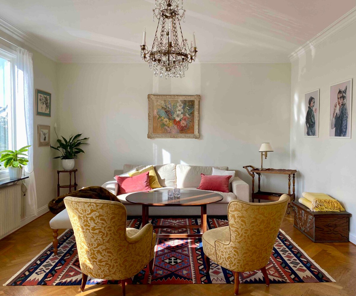 A bright living room is characterized by a soft color palette and elegant décor. Two yellow upholstered chairs frame a central coffee table, while a comfortable sofa offers additional seating. A chandelier hangs from the ceiling, and natural light enters through a large window adorned with white curtains.