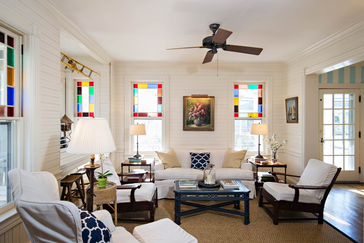 A light-filled living room features white wood paneling and colorful stained glass windows. Comfortable white sofas are arranged around a dark coffee table, complemented by wooden armchairs. A ceiling fan and a pair of table lamps provide additional ambient lighting, enhancing the inviting space.