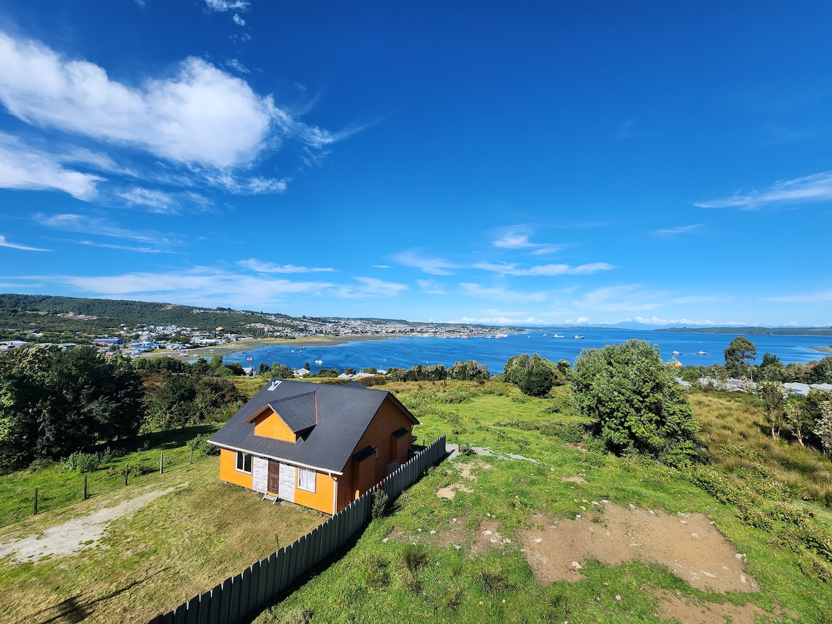 A charming orange house is set against a backdrop of rolling green hills and a vast blue ocean. The structure features a dark roof and large windows, offering a panoramic view of the seascape and surrounding landscapes.