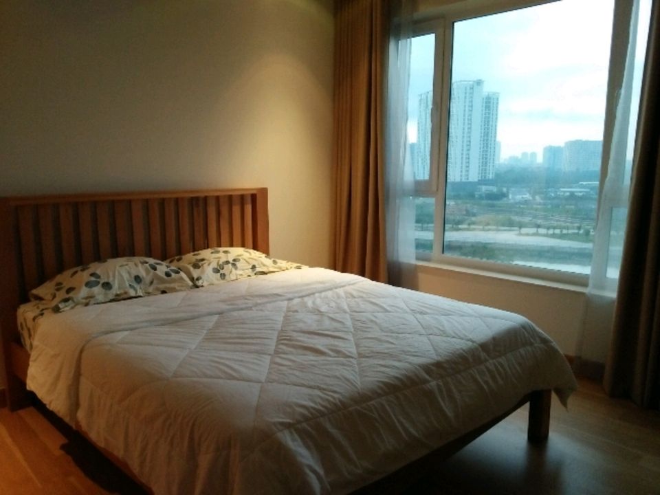 A spacious sleeping area features a wooden bed with a light quilt and patterned pillows. Large window curtains frame the expansive window, allowing natural light to fill the room while offering views of the surrounding cityscape.