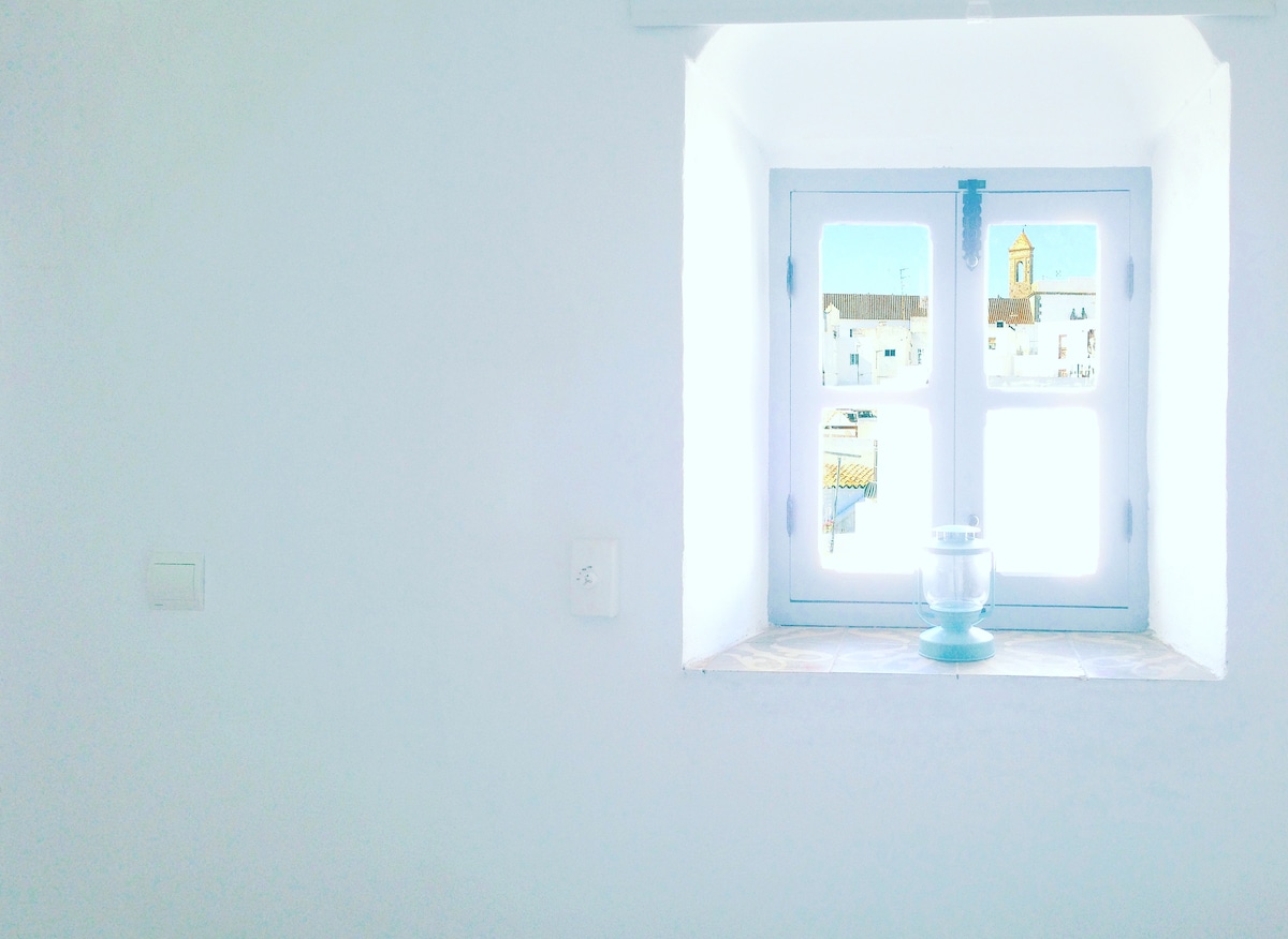 A bright window set within a white wall offers a view of the village. Soft light floods the room, highlighting a simple glass lantern on the windowsill, enhancing the sense of openness and tranquility.