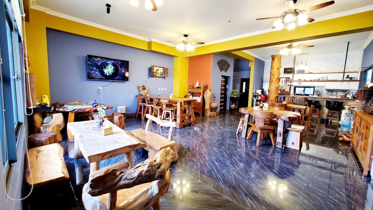 An inviting communal area features natural wood furniture, including various tables and benches. A large television is mounted on the wall, and the kitchen area is visible in the background. Bright colors enhance the warm atmosphere of the space.