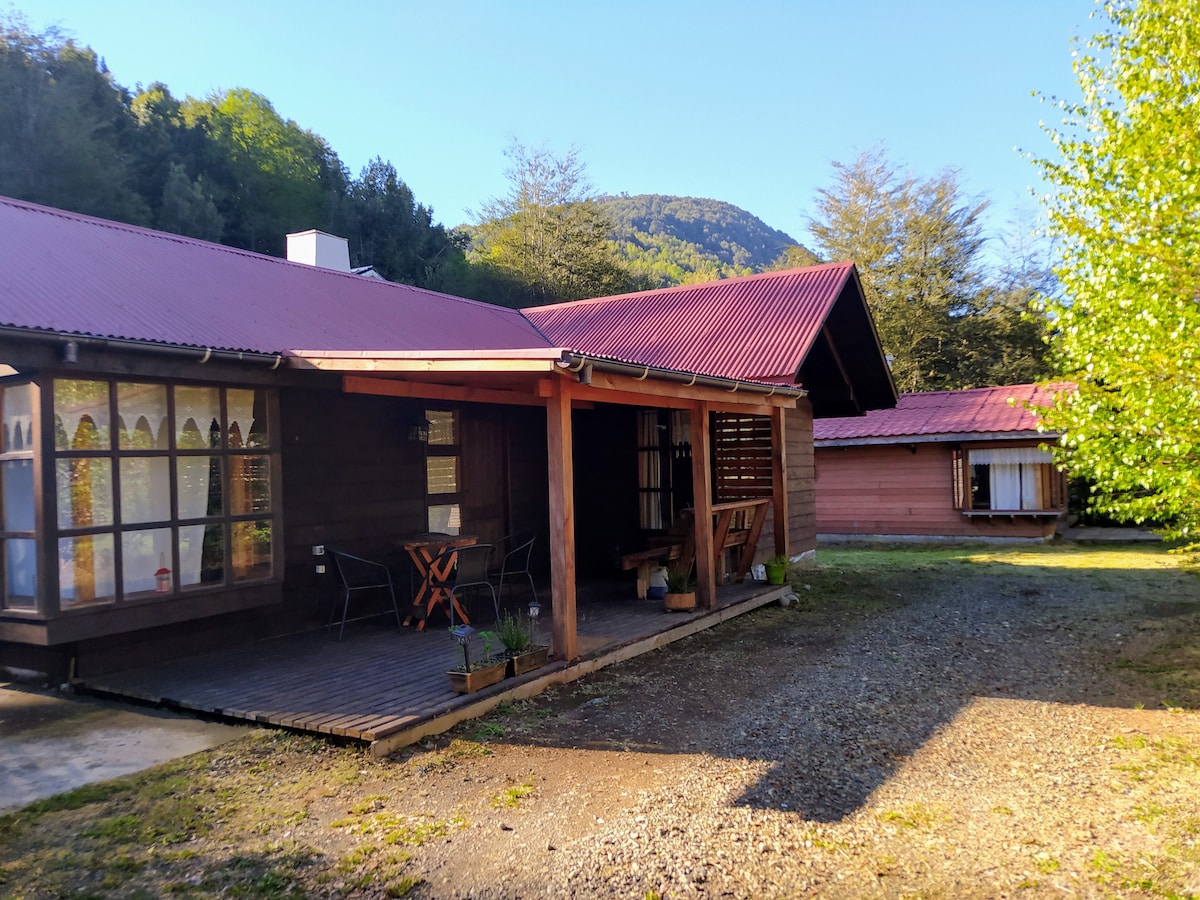 A wooden cabin is featured with a covered porch, two chairs, and a small table on a partially shaded deck. The exterior is complemented by a red metal roof and large windows, providing a view of the surrounding greenery and mountains.