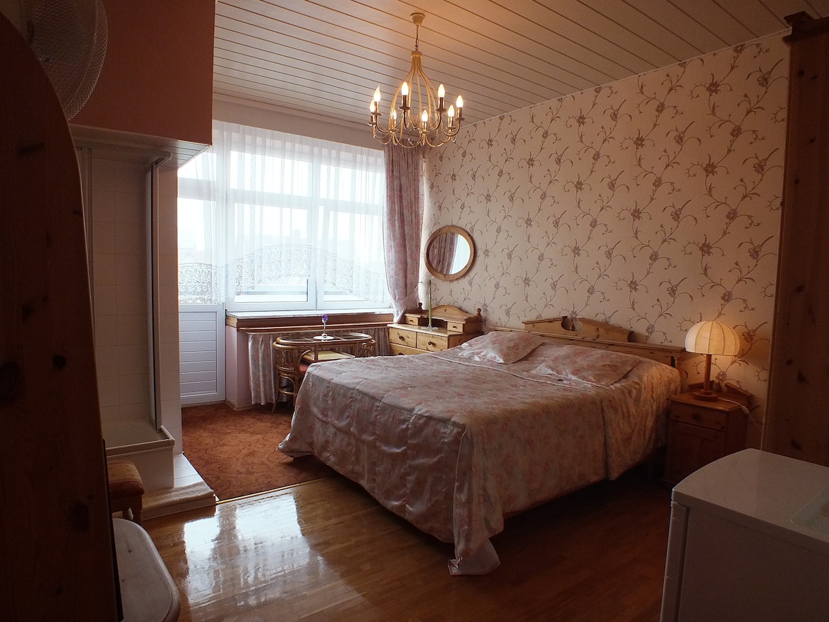 A double room features a spacious bed covered with light floral bedding. Natural light enters through a large window adorned with sheer curtains. Wooden furniture includes nightstands and a desk, complemented by a chandelier and a cozy lamp. A warm carpeted floor adds to the inviting ambiance.