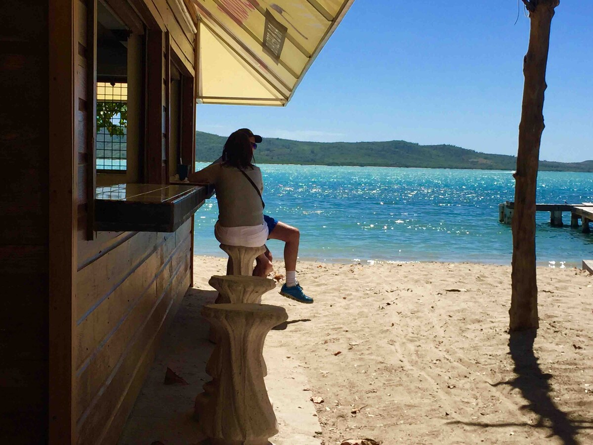 A sandy beachfront is visible, with clear turquoise waters gently lapping at the shore. A person is seated on a stone bench near a wooden structure, enjoying the scenic view of the ocean and distant hills under a bright blue sky.