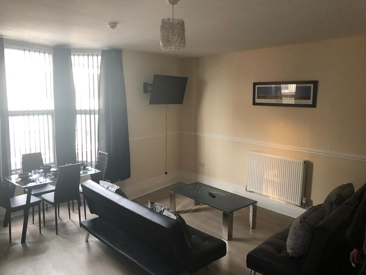 A living area is arranged with a sleek sofa and glass coffee table. Dining space is visible with a table set for four. A wall-mounted television is positioned for viewing, and tall windows adorned with black curtains allow natural light to fill the room.