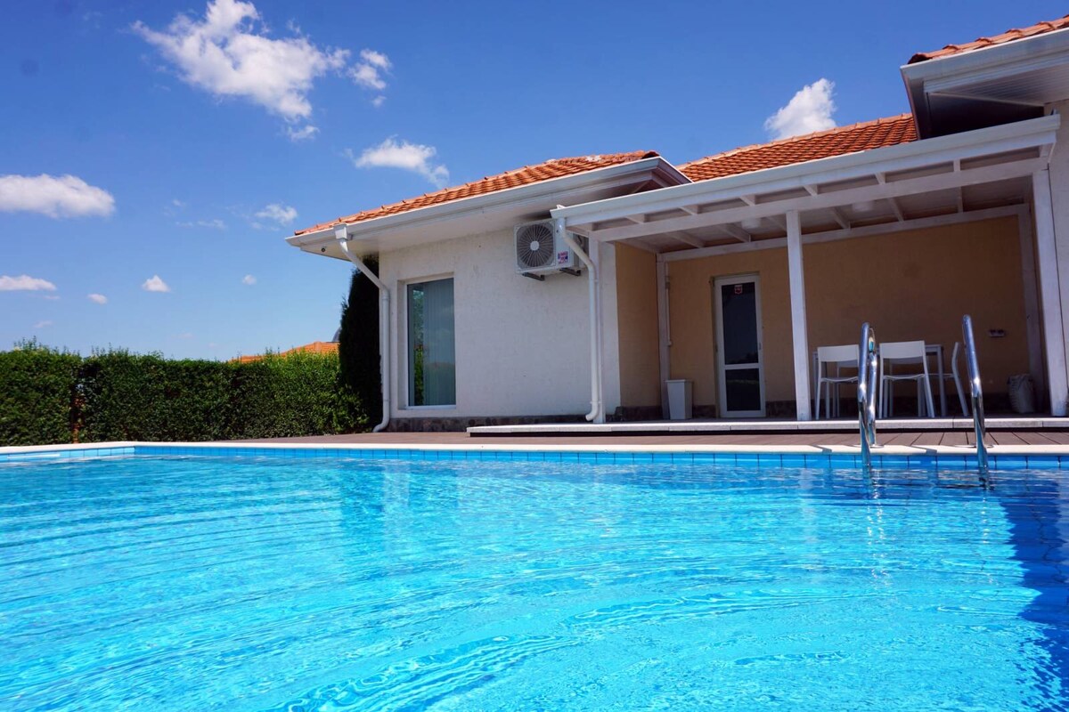 A large swimming pool reflects the clear blue sky, inviting for a refreshing dip. The poolside area features a spacious terrace with seating arrangements visible in the background. The exterior of the guest house is partially shaded by hedges, creating an intimate atmosphere.