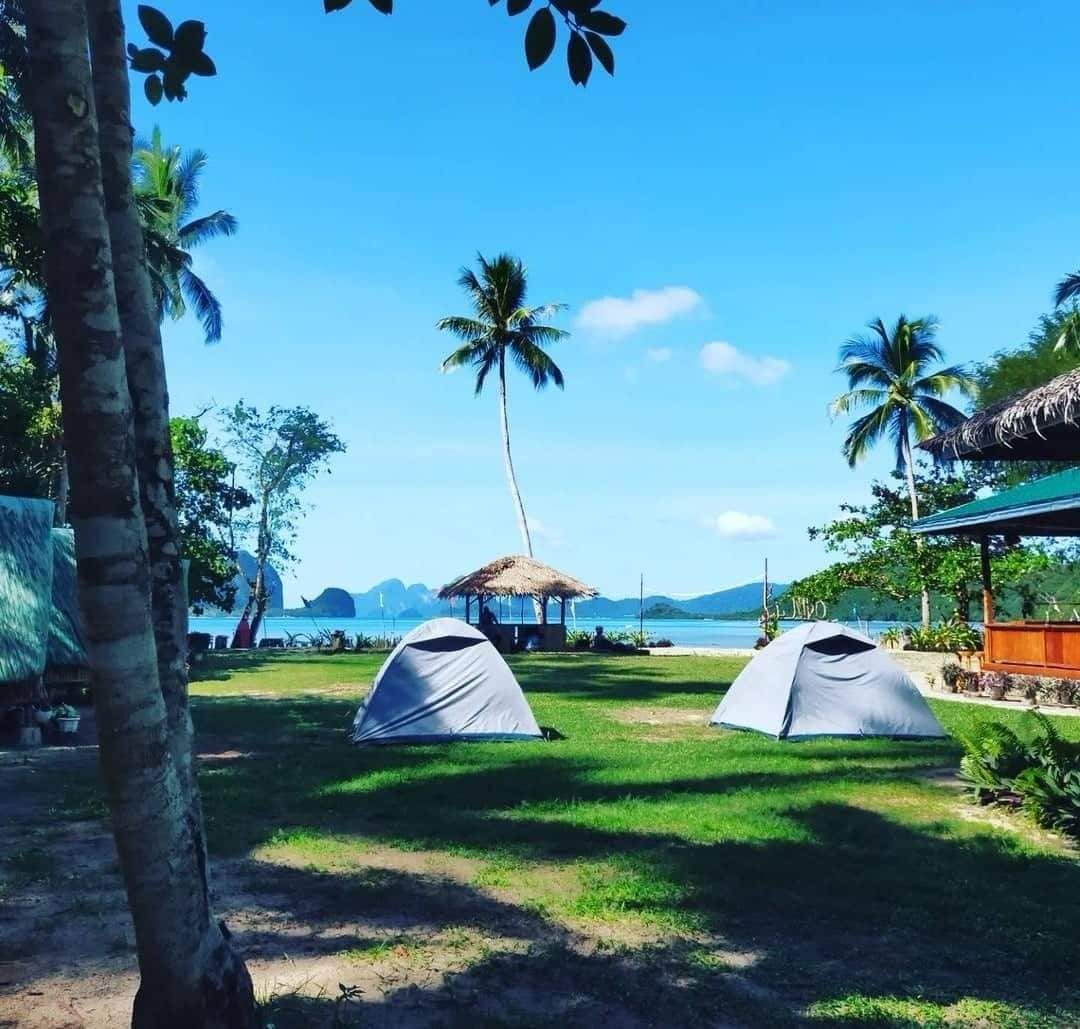Two grey tents are set up on a grassy area, surrounded by palm trees. A thatched-roof gazebo is visible in the background, overlooking a serene beach and distant hills under a clear blue sky.