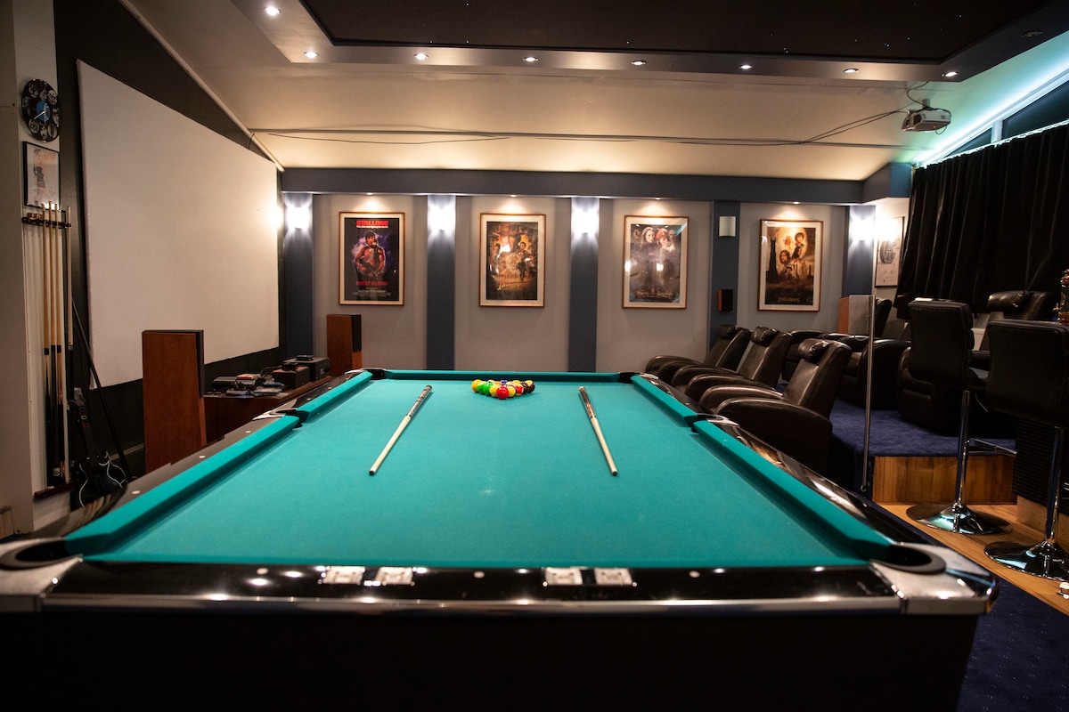 A game room is presented, featuring a well-maintained pool table at center stage. Surrounding the table, leather seating provides a comfortable area for relaxation. Framed artwork adorns the walls, contributing to the room's ambiance. A projector screen is situated at one end of the room.