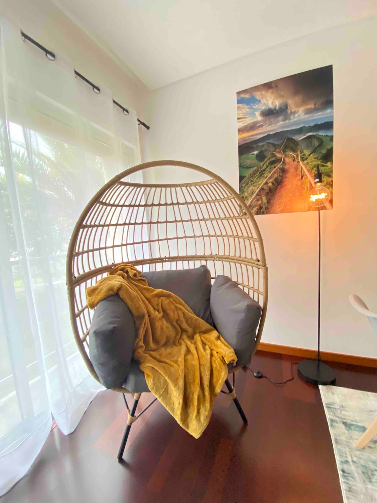 A unique egg-shaped chair, with plush grey cushions, is positioned in a well-lit corner. A warm-colored throw is draped over the chair. A floor lamp emits soft light, and a scenic landscape photograph hangs on the wall, complementing the serene ambiance.