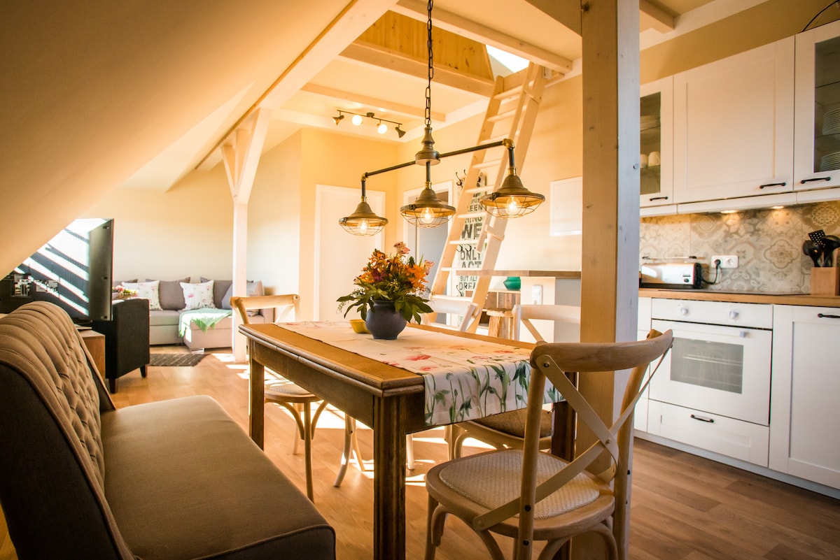 A well-lit kitchen and dining area features a wooden dining table surrounded by chairs. Overhead lights provide illumination. A comfortable sofa is present in the background, and a ladder leads to a lofted space. The kitchen includes modern appliances and cabinetry.