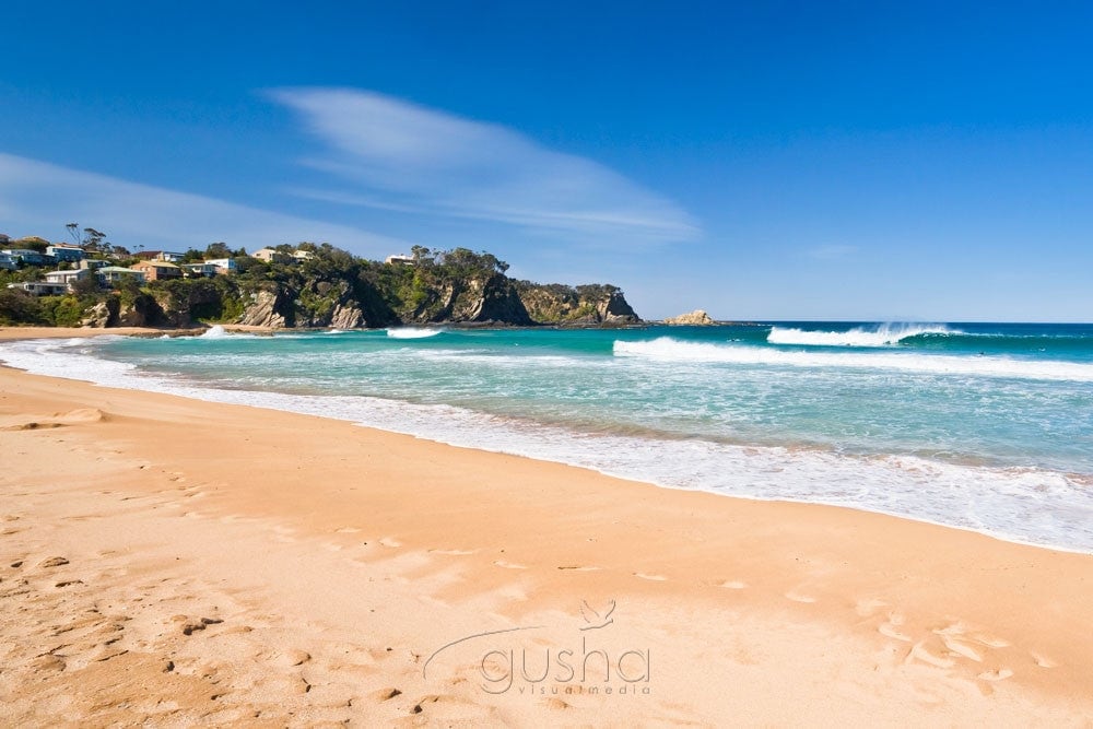 A serene beach scene is captured, featuring soft golden sand and gentle waves lapping at the shoreline. In the distance, rugged cliffs meet the ocean under a clear blue sky, with a few coastal homes visible among the trees.