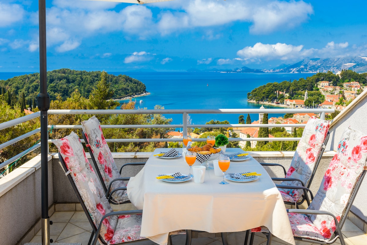 A balcony table set for breakfast is presented, featuring fruit and drinks against a backdrop of the sea and horizon. Surrounding chairs, adorned with floral fabric, overlook lush greenery and colorful coastal buildings, inviting moments of relaxation and enjoyment in a scenic setting.