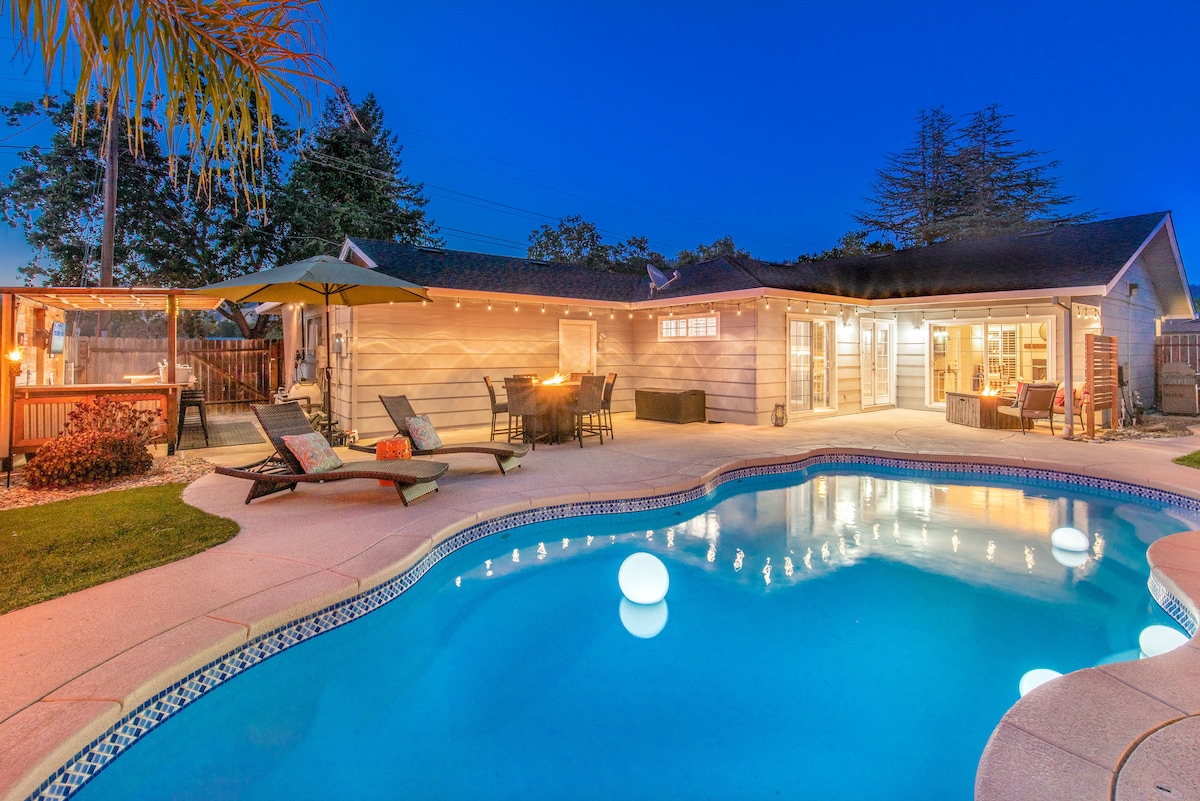 An inviting outdoor area features a heated pool with illuminated spheres floating on the surface. Comfortable loungers are arranged nearby. A dining set with a fire pit stands under a shaded pergola, creating a suitable space for outdoor gatherings. The home is softly lit in the background.