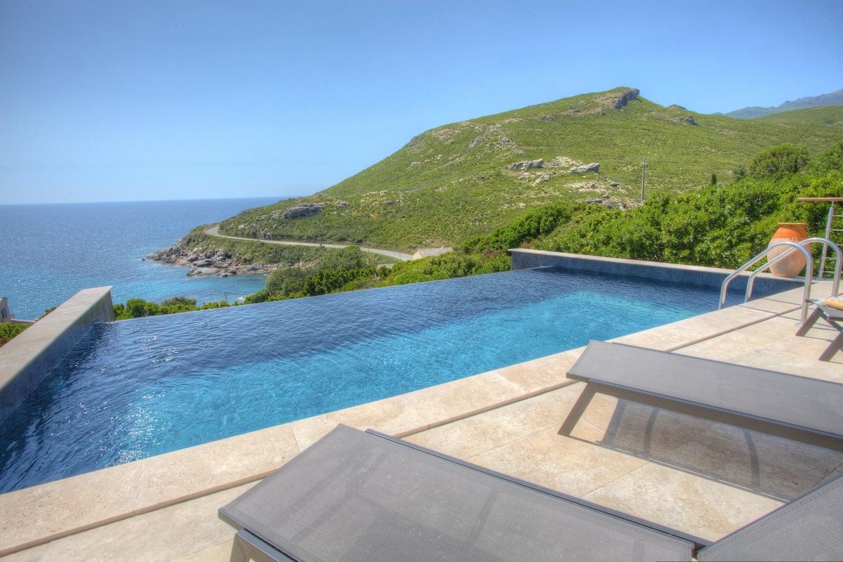 An infinity pool extends towards the horizon, offering panoramic views of the coastline and surrounding mountains. Lounge chairs are positioned along the pool's edge, providing a relaxing space to enjoy the scenic vista. The clear sky enhances the tranquil atmosphere of the landscape.