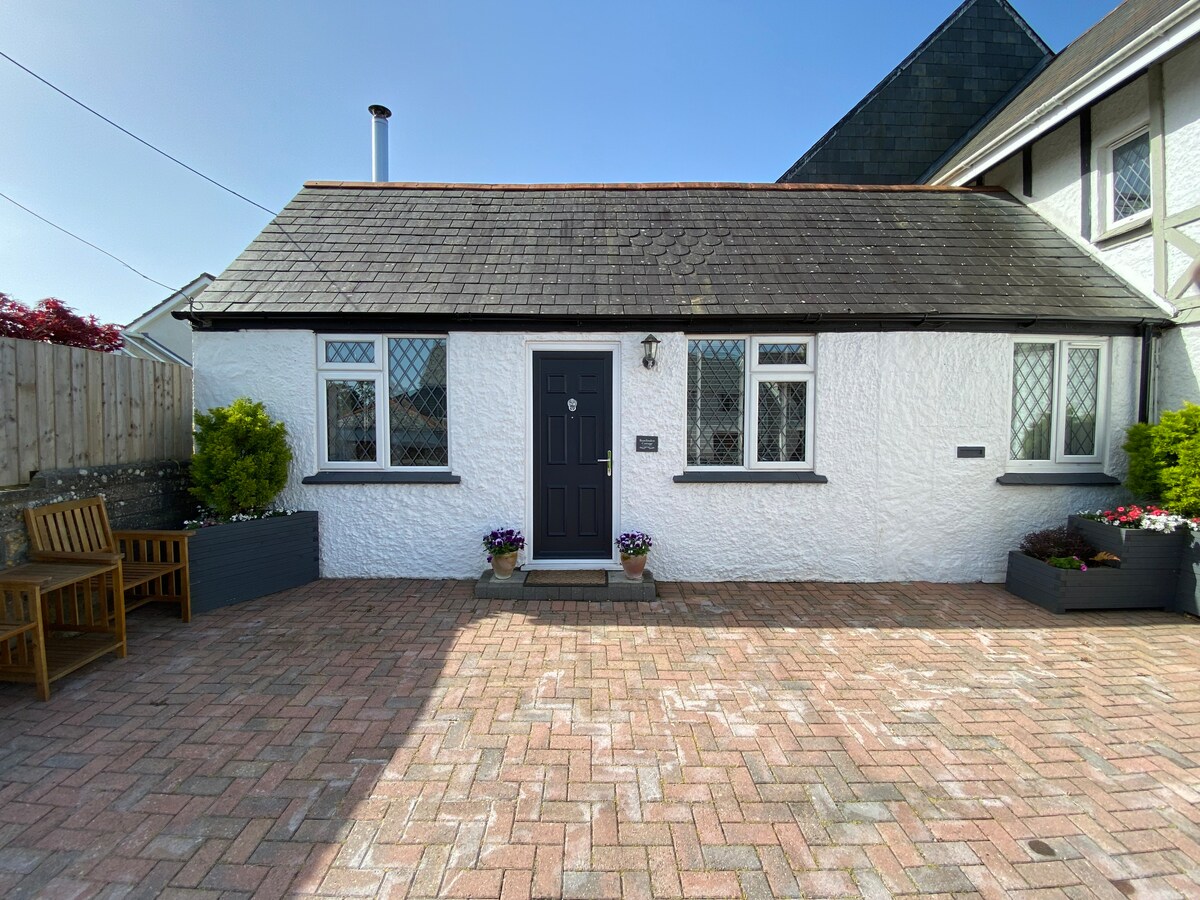 The exterior of Roselinden Cottage is depicted, showcasing a charming whitewashed façade with a black front door. Flanking the entrance are potted plants, and outdoor seating is available on a brick-paved area, offering a welcoming first impression.
