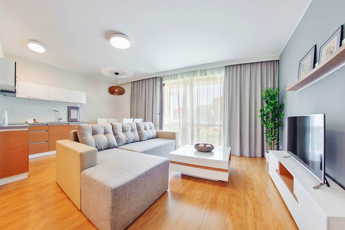 A modern living area is displayed, featuring a large sectional sofa and a coffee table. Natural light filters through sheer curtains, illuminating the space. An open kitchen with sleek cabinetry is visible in the background, complemented by a decorative plant and a flat-screen television.