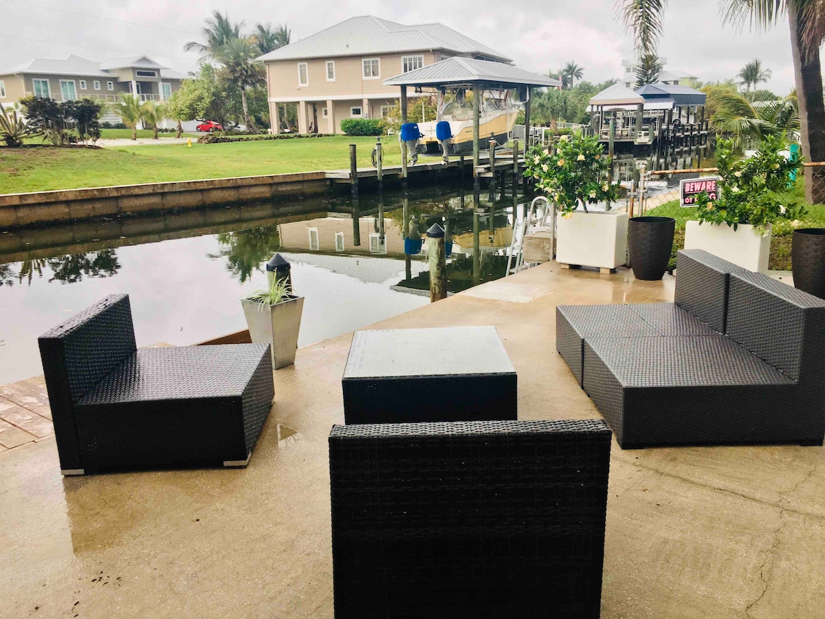 An outdoor seating area is set beside a calm canal. Dark wicker furniture, including a table and several modular chairs, is arranged on a concrete surface. Lush greenery is visible in planters, along with reflections of nearby homes on the water's surface.