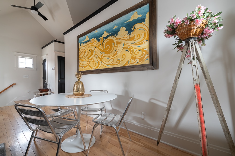 A round dining table is set with a woven placemat, surrounded by four metallic chairs. A large piece of colorful wall art depicting abstract waves dominates the space, while a decorative ladder holding a basket of flowers adds a touch of charm.