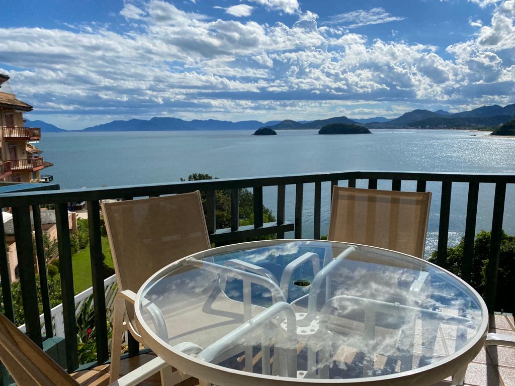 A round glass table with four chairs is positioned on a balcony overlooking the ocean. The view features distant islands and a vibrant sky filled with clouds. The serene waters reflect the surrounding landscape, providing a tranquil setting for outdoor relaxation.