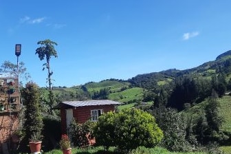 The image presents a scenic countryside view featuring rolling hills and greenery under a clear blue sky. A small wooden cabin is positioned amidst lush vegetation, surrounded by potted plants. The landscape conveys a sense of tranquility and connection to nature.