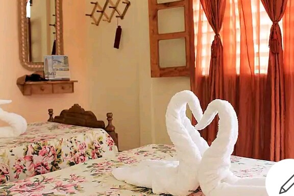 A guest room features two beds adorned with floral bedding. Towels are elegantly arranged in the shape of swans on the beds. Natural light filters through sheer curtains, casting a warm glow. A wooden mirror and wall hooks add functional and decorative elements to the space.