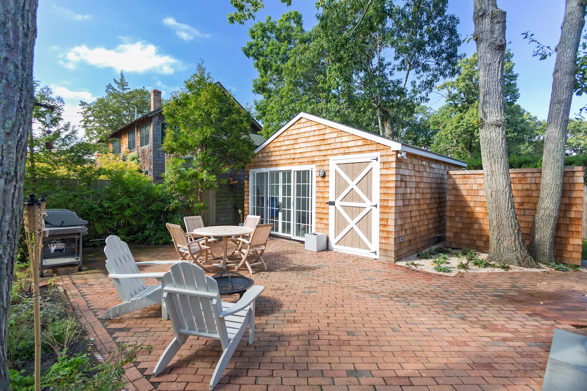 A spacious outdoor patio is surrounded by trees, featuring a circular table with four chairs. Two adirondack chairs are positioned on a brick surface, creating a relaxed seating area. A grill is located nearby, and a shed with white doors adds charm to the setting.