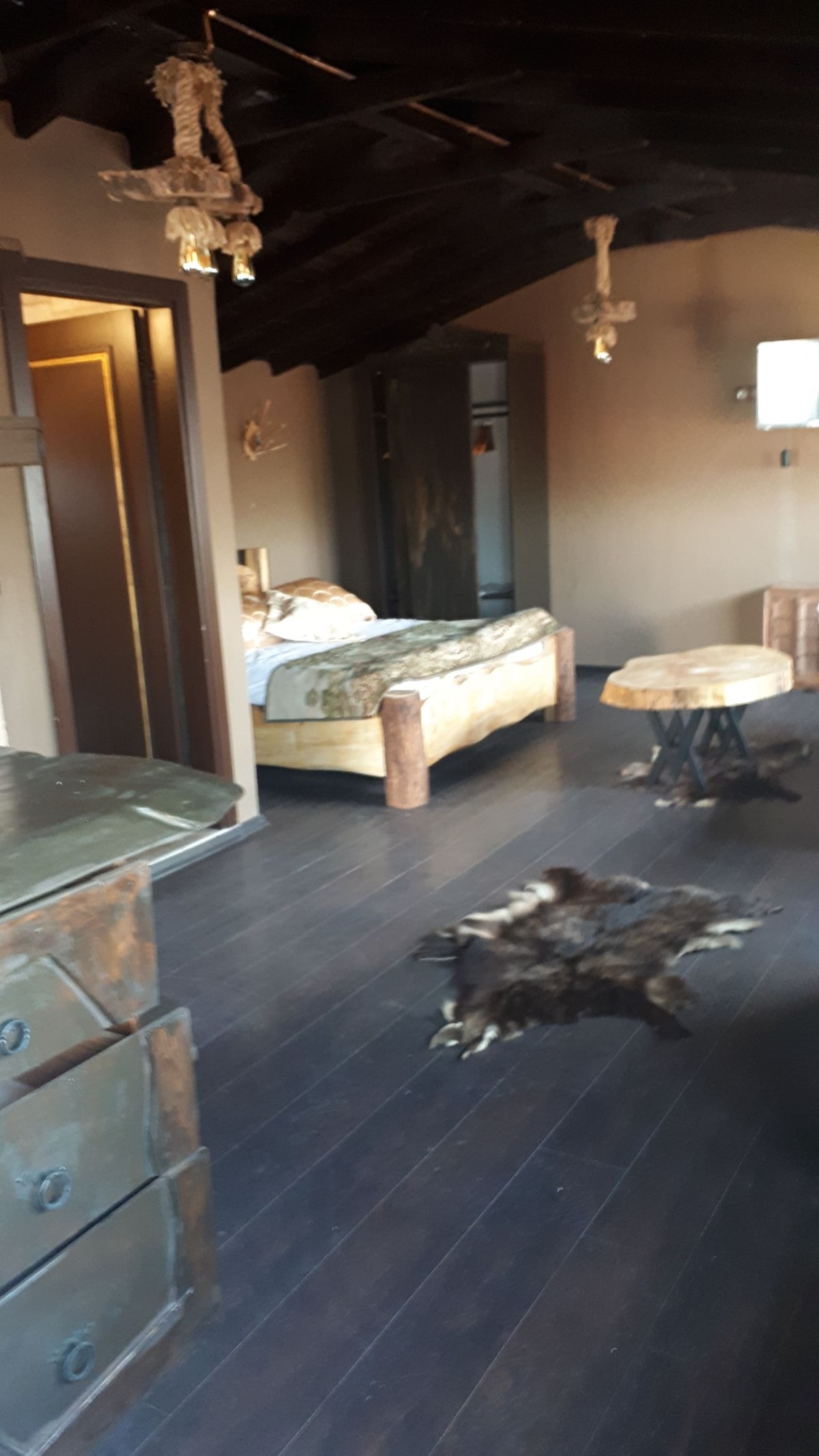 A spacious bedroom features a wooden bed with decorative bedding. A rustic wooden table sits near a cowhide rug on dark wooden flooring. A television is mounted on the wall, with a closet visible in the background, adding to the room's functional design.