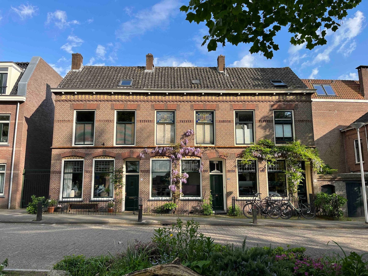 A historic brick building displays distinct architectural features, with four large front windows framed by decorative elements. A vibrant wisteria plant climbs the facade, adding color. Bicycles are parked in front, with a landscaped area showcasing green plants along the pathway.