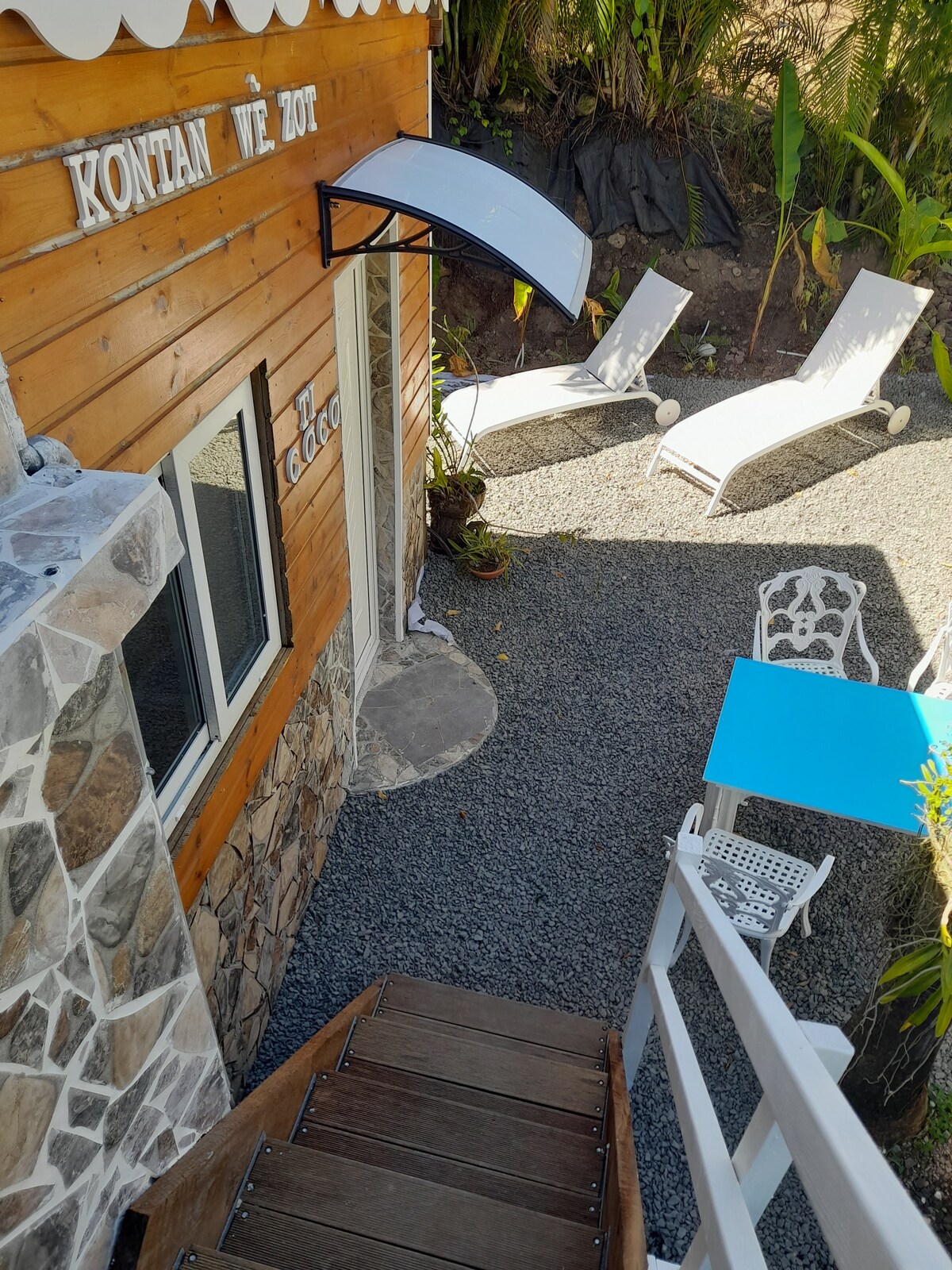 An outdoor area is visible, featuring lounge chairs and a small table set on gravel. The steps lead down from the wooden entrance, framed by natural greenery and stone accents. A bright blue table adds a pop of color, inviting relaxation in this serene environment.