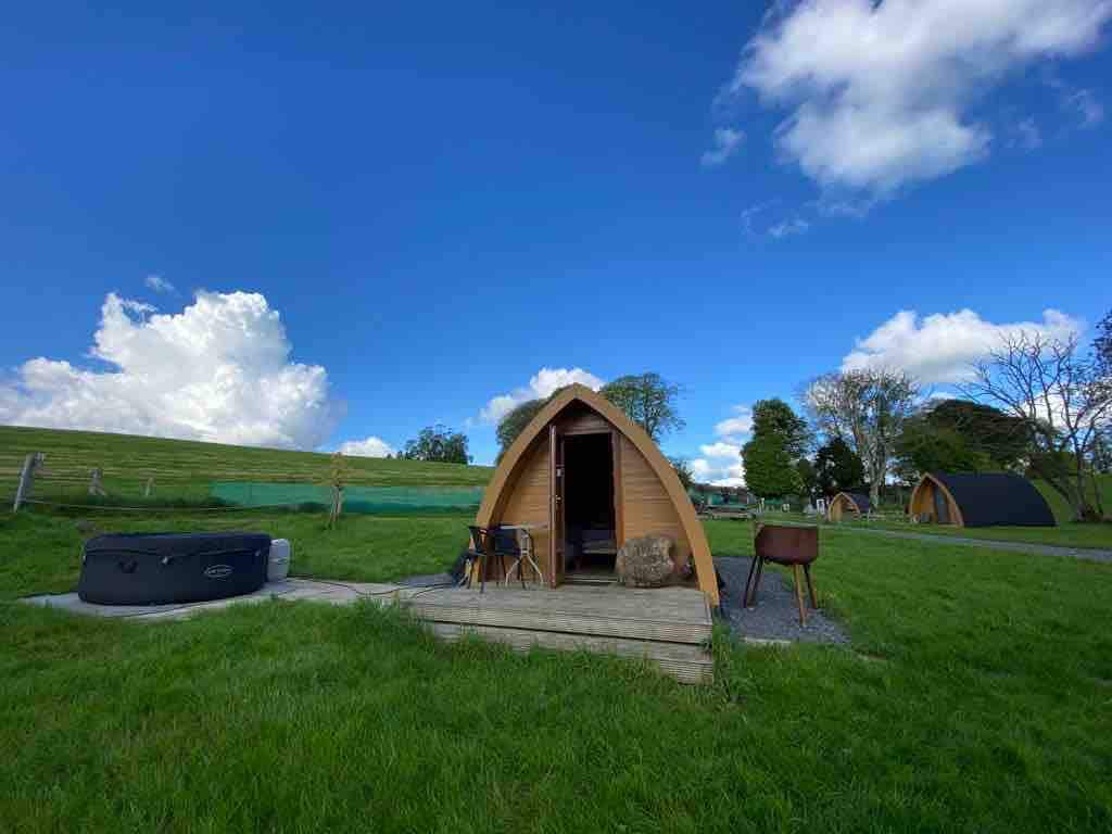 A glamping pod with a pointed roof is visible, situated on a grassy area. A wooden deck leads to the entrance. A hot tub is positioned nearby, surrounded by green space. The backdrop features a clear blue sky with fluffy clouds.