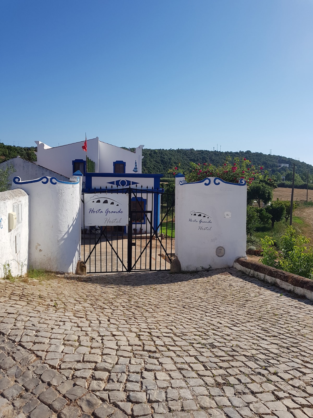 The entrance to Horta Grande Hostel is marked by a decorative white wall with blue accents. A black gate provides entry, framed by lush greenery and flowers. Rolling hills can be seen in the background under a clear blue sky, enhancing the outdoor ambiance.