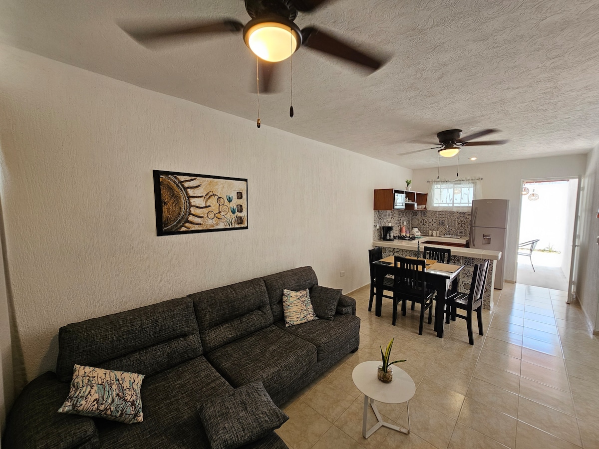 A spacious living area is furnished with a dark sectional sofa, accented by decorative pillows. A small table sits in front of the sofa. A dining table with black chairs is visible nearby, with a modern kitchen in the background featuring wooden cabinetry and a neutral tile floor.