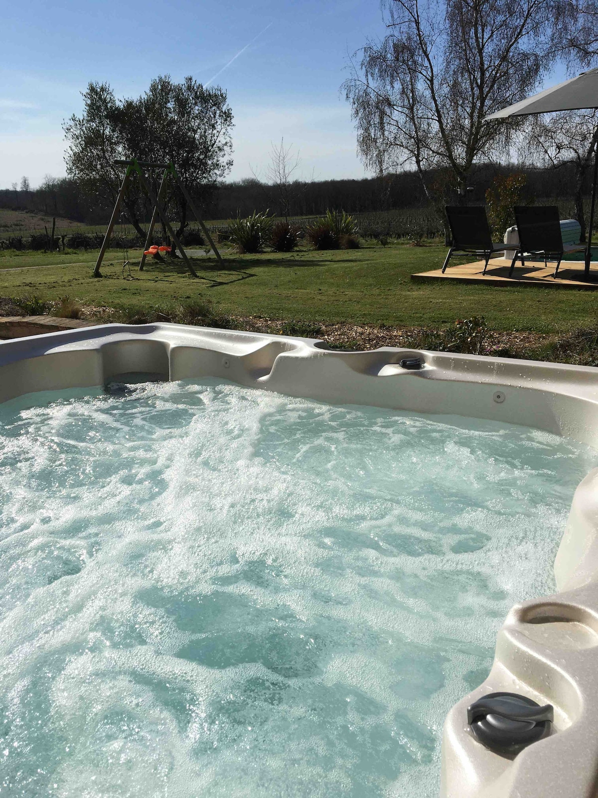 A private spa with bubbling water is set in a serene outdoor area, surrounded by lush greenery. In the background, a swing set and relaxation area with outdoor seating can be seen, offering a peaceful view of the landscape.