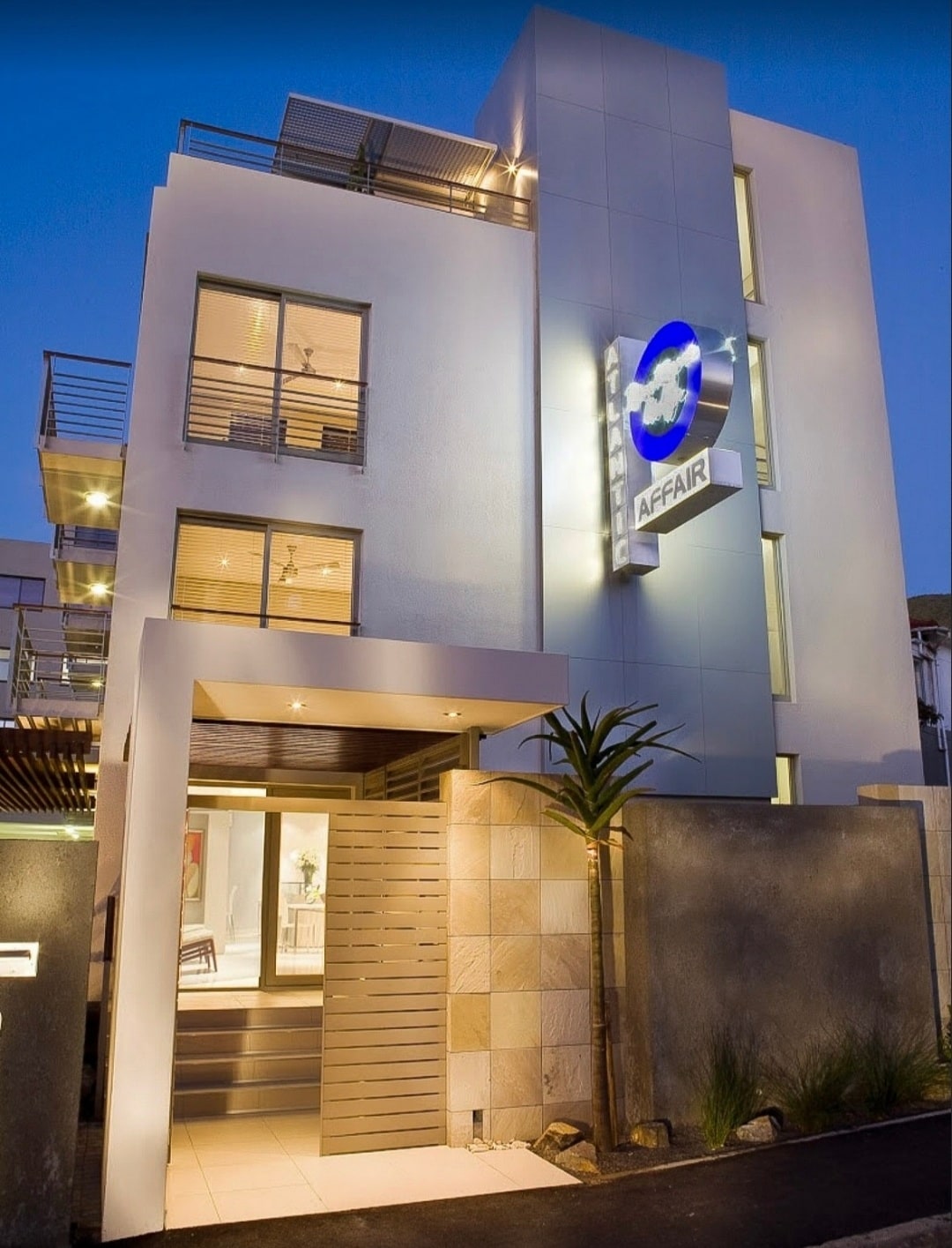The modern exterior of the hotel is featured, showcasing sleek lines and a minimalist design. The entrance is framed by a decorative stone wall and illuminated signage. Large windows on the upper levels reflect the evening light, signaling a welcoming atmosphere.