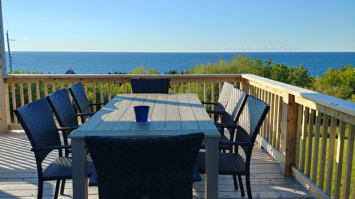 An outdoor deck is presented with a large wooden table surrounded by six woven chairs. A small blue pot is centered on the table, while an unobstructed view of the ocean is seen in the distance, complemented by a clear blue sky above.