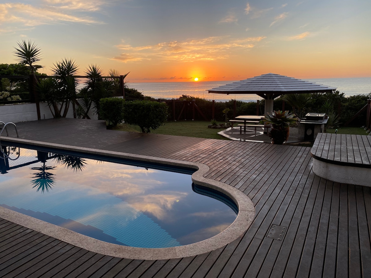 A serene view of a swimming pool reflects the vibrant colors of a sunset over the ocean. Surrounding greenery and a shaded barbeque area enhance the outdoor space, creating a peaceful atmosphere for relaxation.