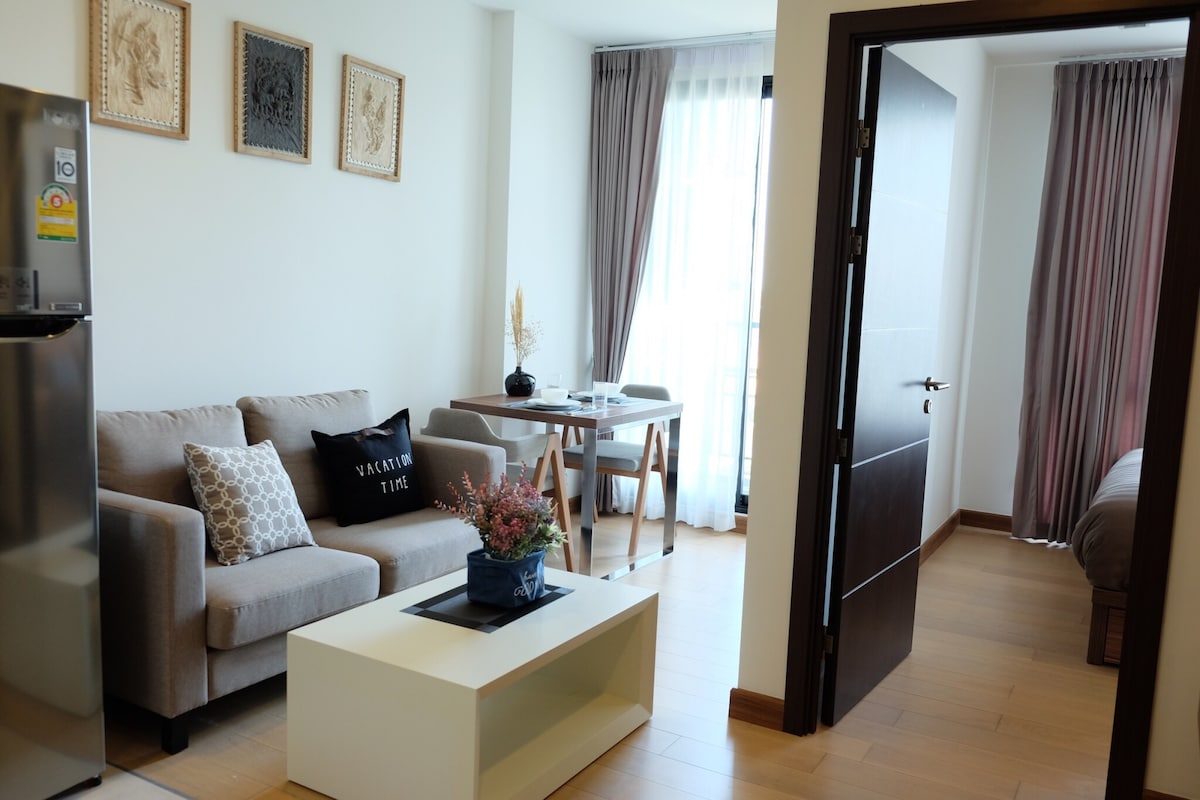 The living area features a comfortable sofa and a coffee table, with decorative cushions adding a touch of detail. A dining table is positioned near a window, enhancing the natural light. A doorway leads to the bedroom, offering a glimpse of the warm, inviting interior.