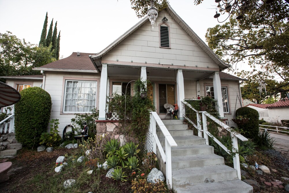 The exterior of a craftsman-style house is depicted, featuring a large staircase leading to a welcoming front porch. Lush greenery and decorative plants are arranged around the entrance, while tall trees stand in the background, contributing to a serene setting.