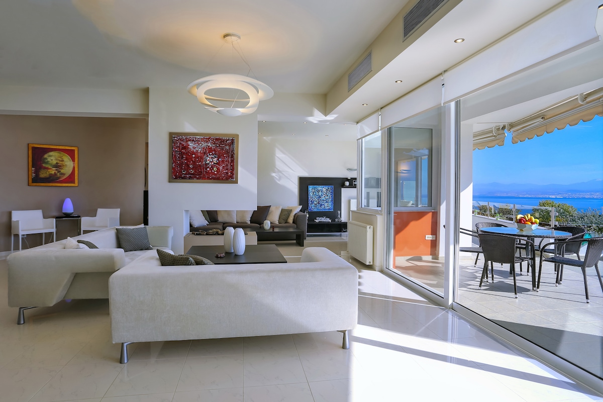 A spacious living area is highlighted by large glass doors, connecting to an outdoor dining space with panoramic views. A modern sectional sofa and coffee table are arranged around a decorative wall art piece, with a contemporary light fixture hanging above.