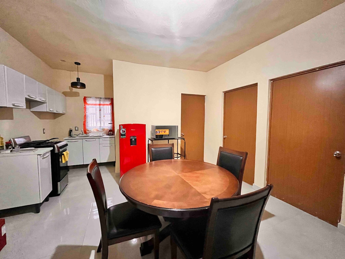 A bright kitchen area features white cabinets and appliances, including a red refrigerator. A wooden dining table is surrounded by four black chairs. Two doors provide access to additional spaces, while the overall layout suggests a functional and inviting environment.
