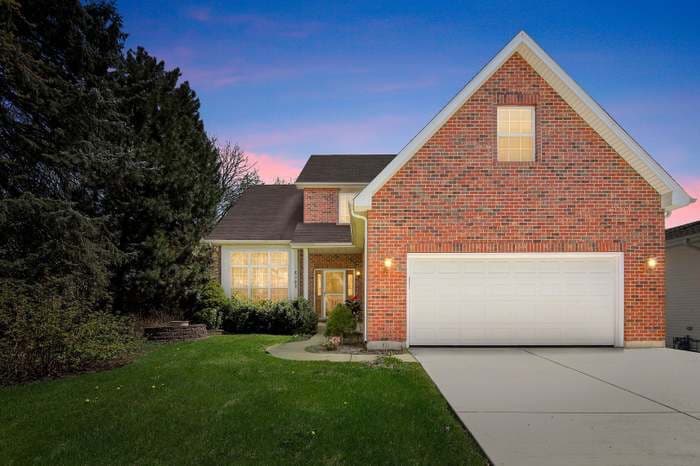 A two-story brick house is positioned among greenery, with well-manicured lawns. Warm light emanates from the front windows, creating a welcoming ambiance. The driveway leads to a spacious garage, and the exterior features a mix of shrubbery and trees, enhancing the property's charm.