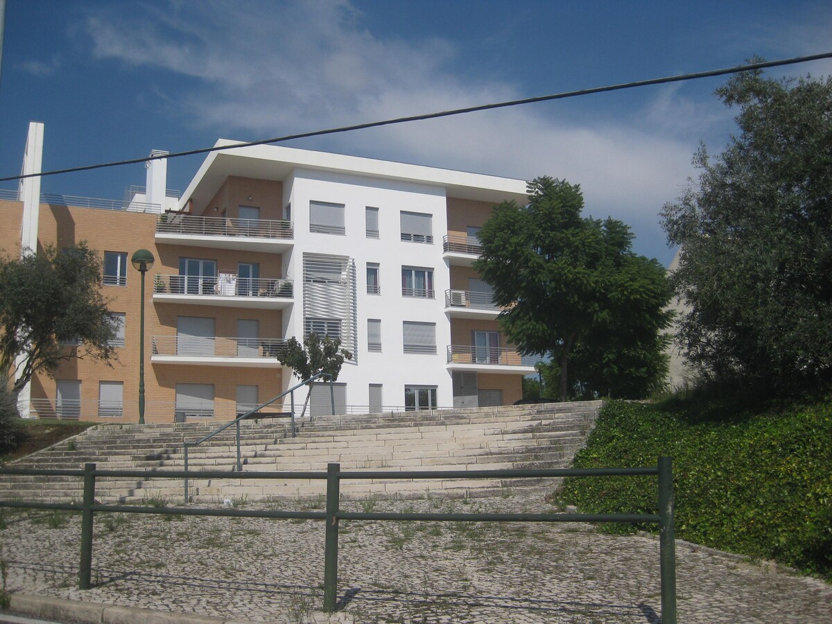 A modern three-story apartment building is presented, featuring a combination of light-colored and white facades. Green trees and shrubs frame the property, while stairs lead to the entrance. Large windows are visible, with some balconies extending from the upper floors.