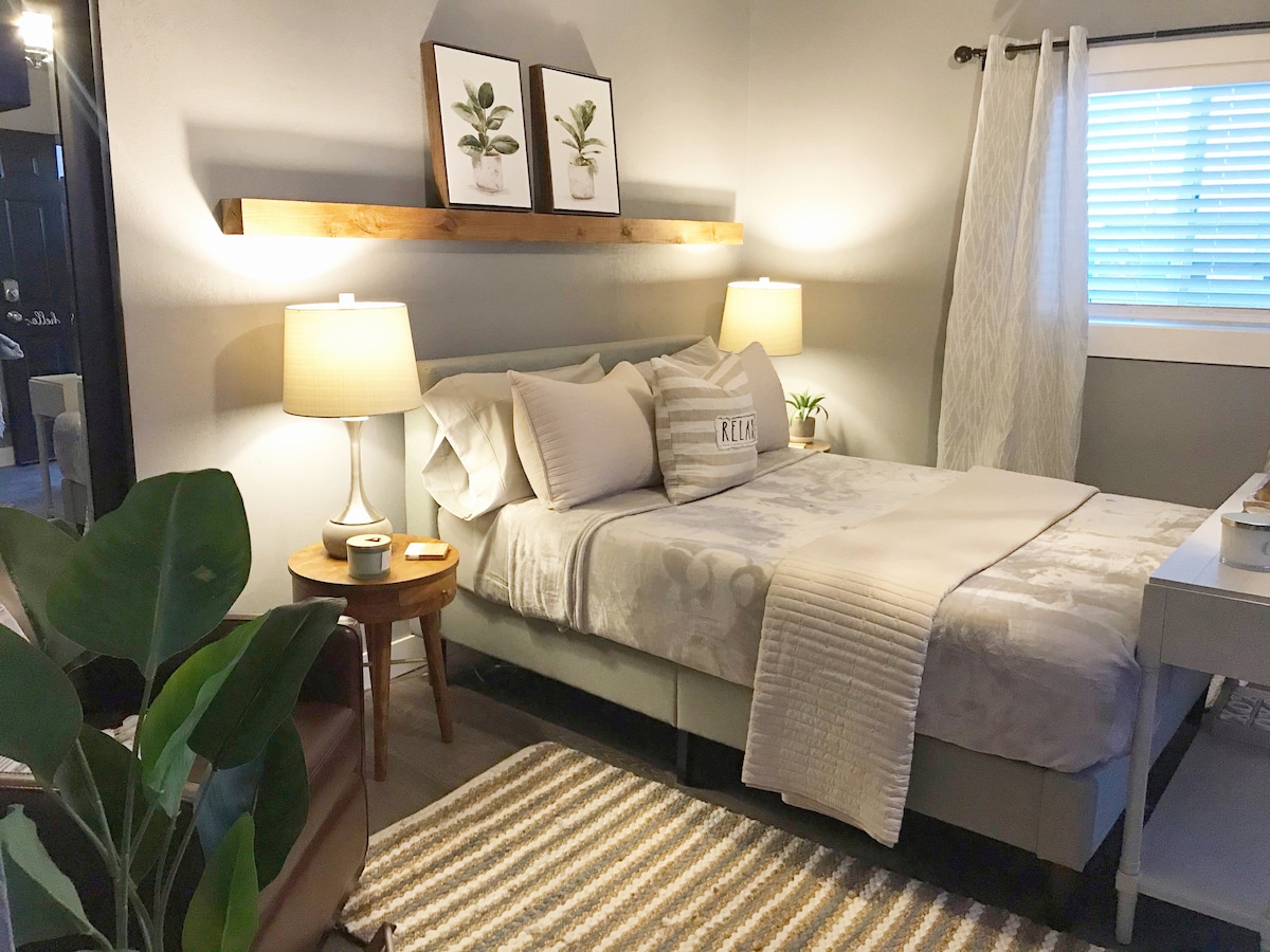 A cozy sleeping area features a spacious bed with neutral bedding and decorative pillows. Two bedside lamps illuminate the space, while a wooden shelf holds framed plant art above the bed. A small plant sits on a nightstand, enhancing the calming ambiance.